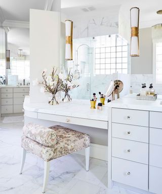 Large white bathroom space with enhanced vanity area, seating and cabinet space. Large mirror with two matching wall lights, marble wall and floor tiles.