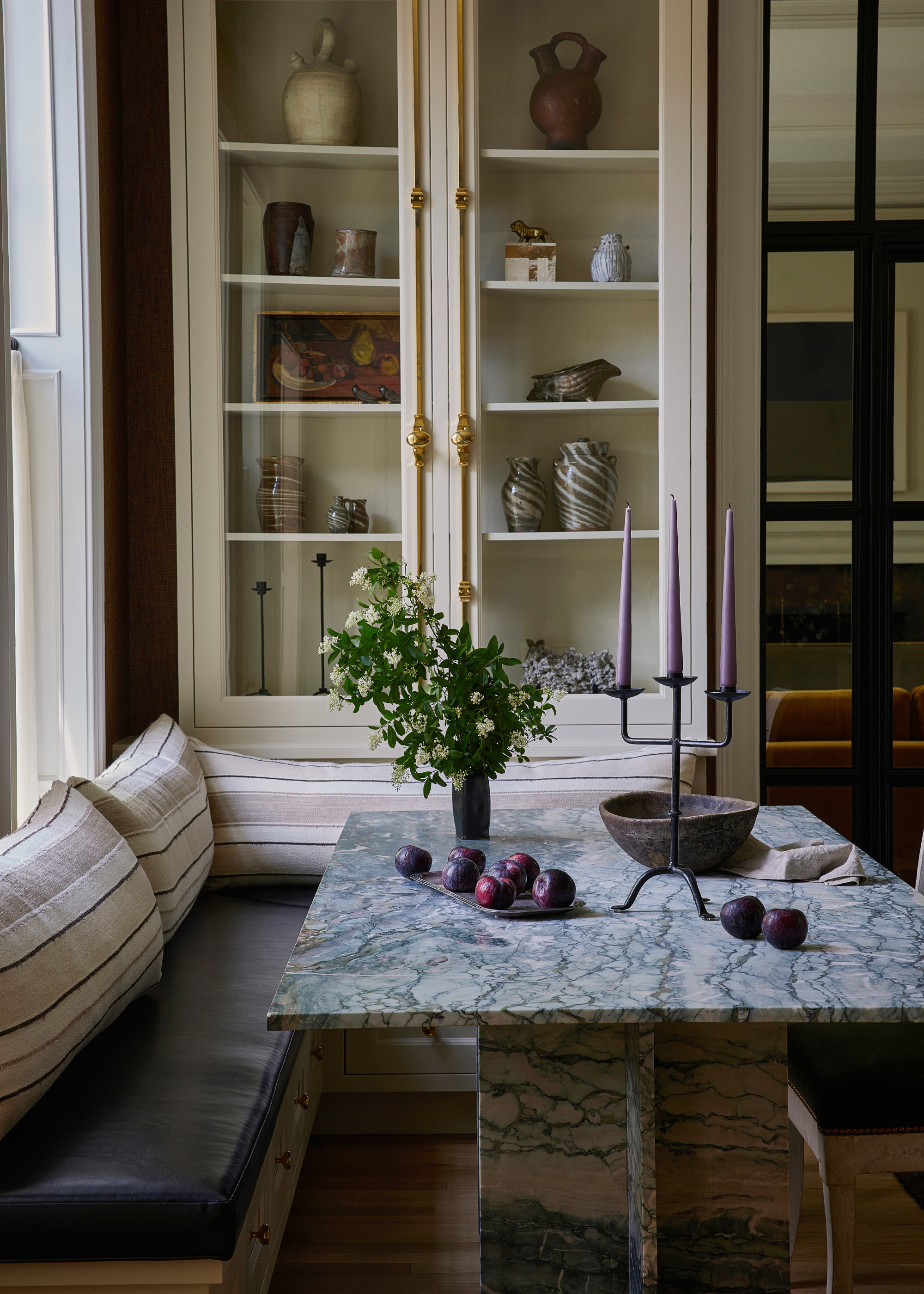 Banquette corner seating with black leather seat cushions and striped back cushions around a green marble table with a plant, candelabra and bowl on top with plums scattered across it with glass-fronted display cabinets behind