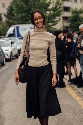 a guest at milan spring/summer 2025 fashion week wears a turtleneck and black midi skirt and glasses