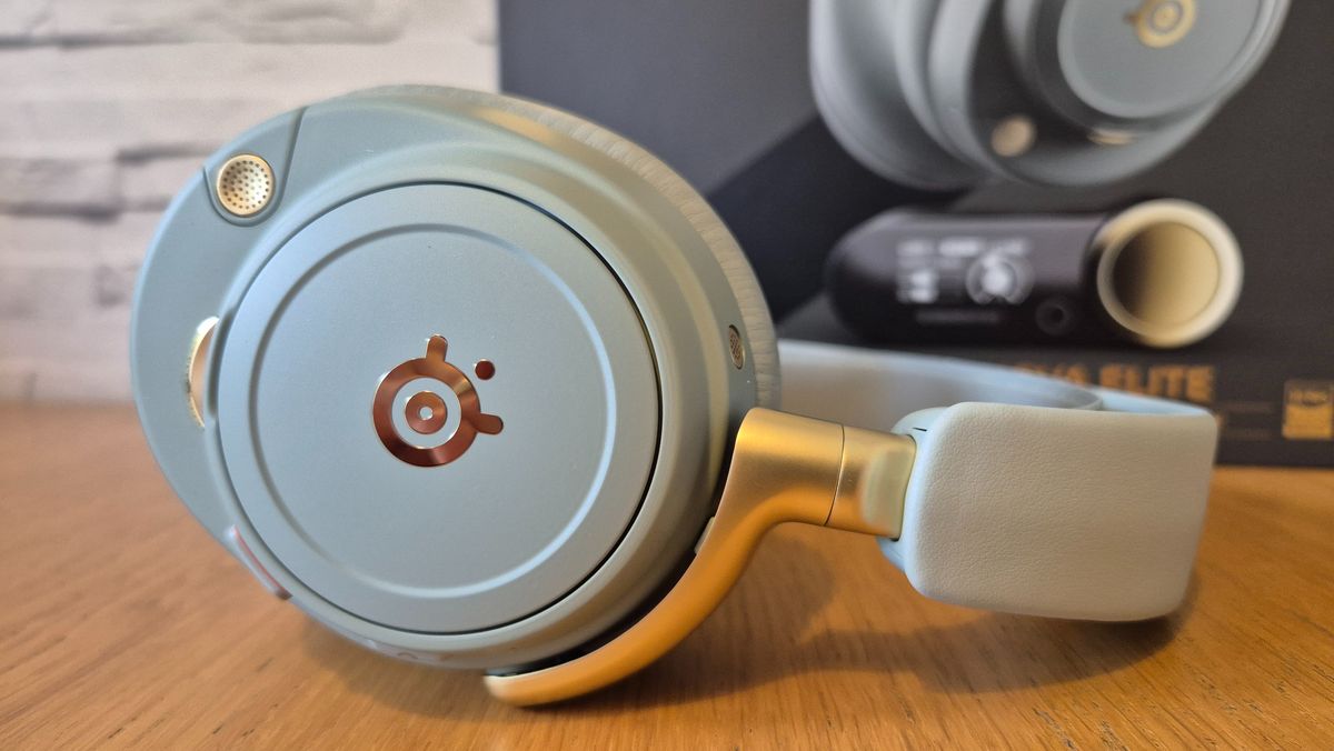The SteelSeries Arctis Nova Elite headset in front of a white brick background and on a wooden surface