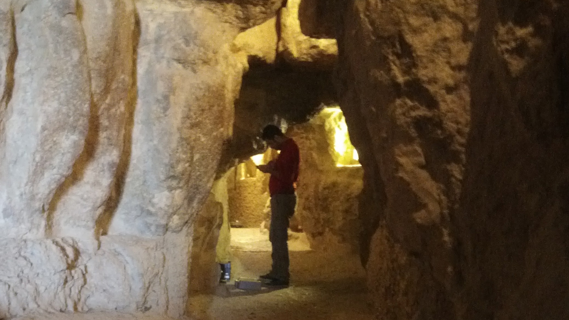 A man wearing a red jacket and dark pants stands in a stone tunnel that's dimly lit.