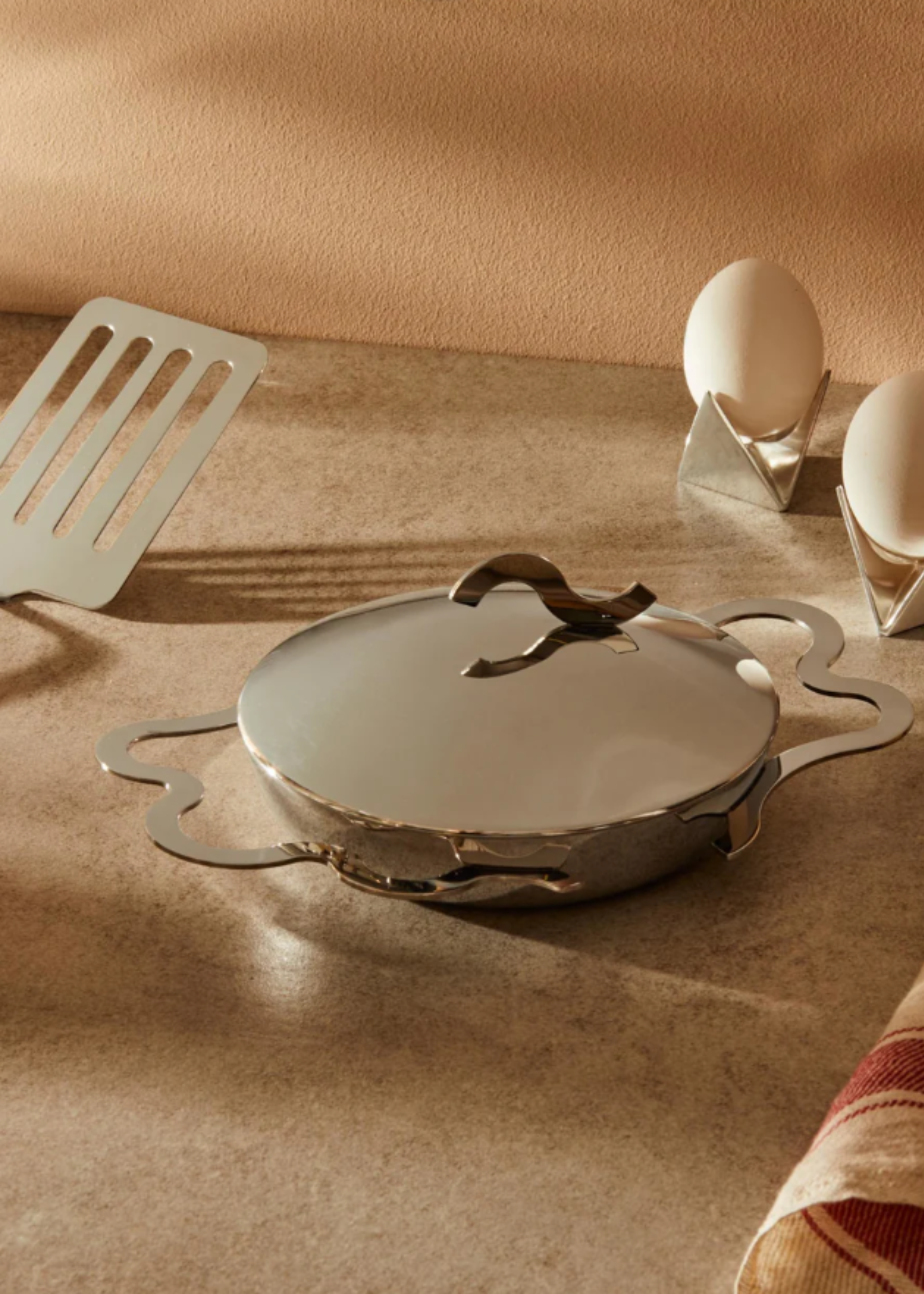 A silver-colored egg frying pan with curved handles and a lid near a slotted turner and eggs on egg cups