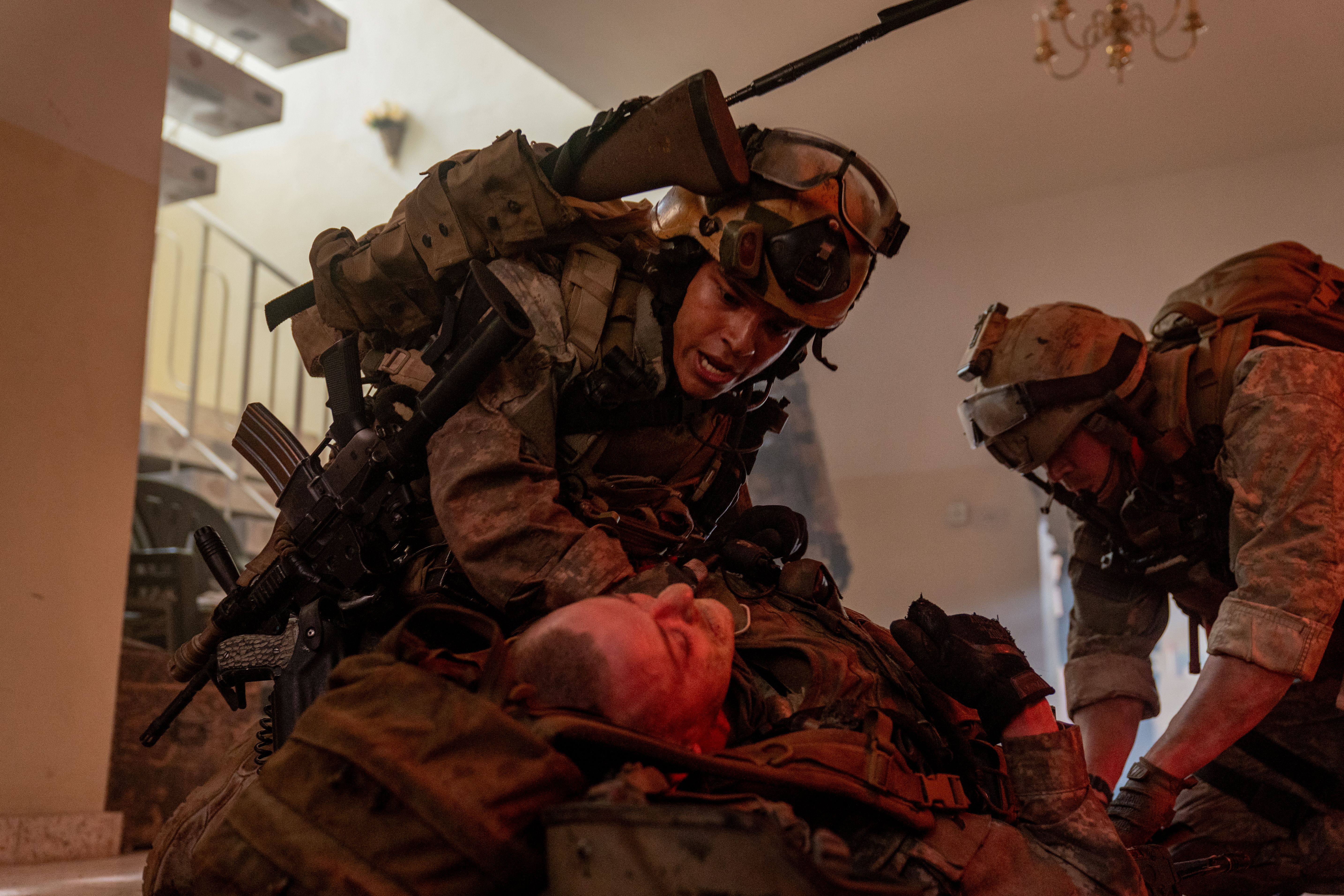 Two soldiers in full gear kneeling to tend to an injured solider on the ground.