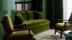 Close-up image of a living room with glossy, dark green walls. There is a velvet, olive green sofa and two velvet, green accent chairs.