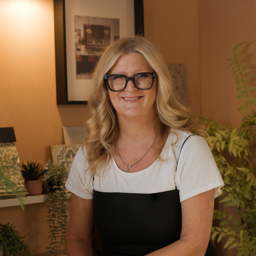 female with long blond hair, wearing glasses, white top and black dress sat in room with pale terracotta walls, decorated with artwork and plants on a shelf