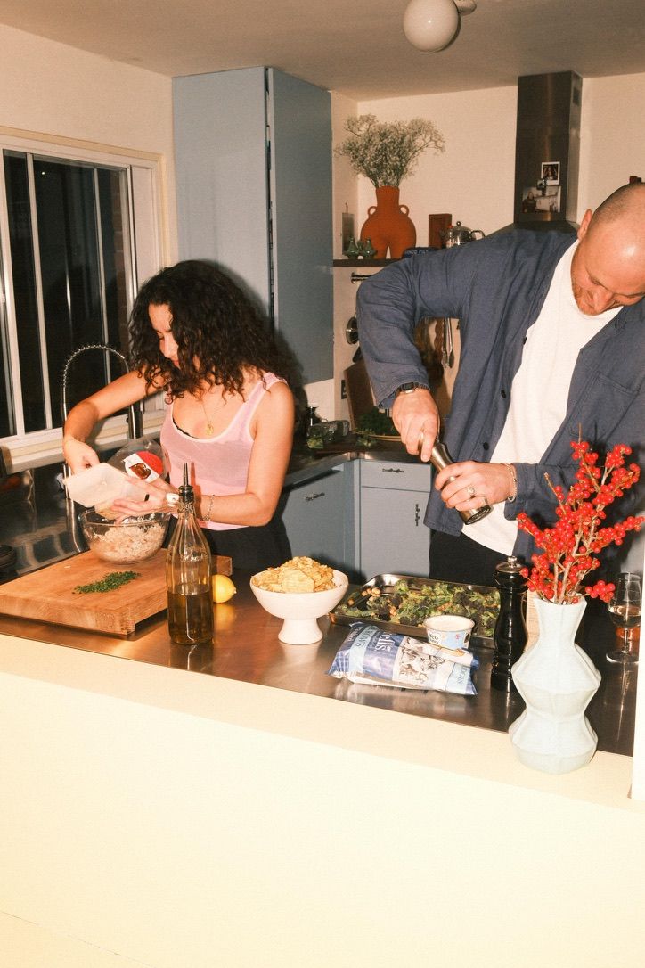 Image of two people cookin in a small kitchen that has light blue cabinetry, stainless steel countertops, and white walls.