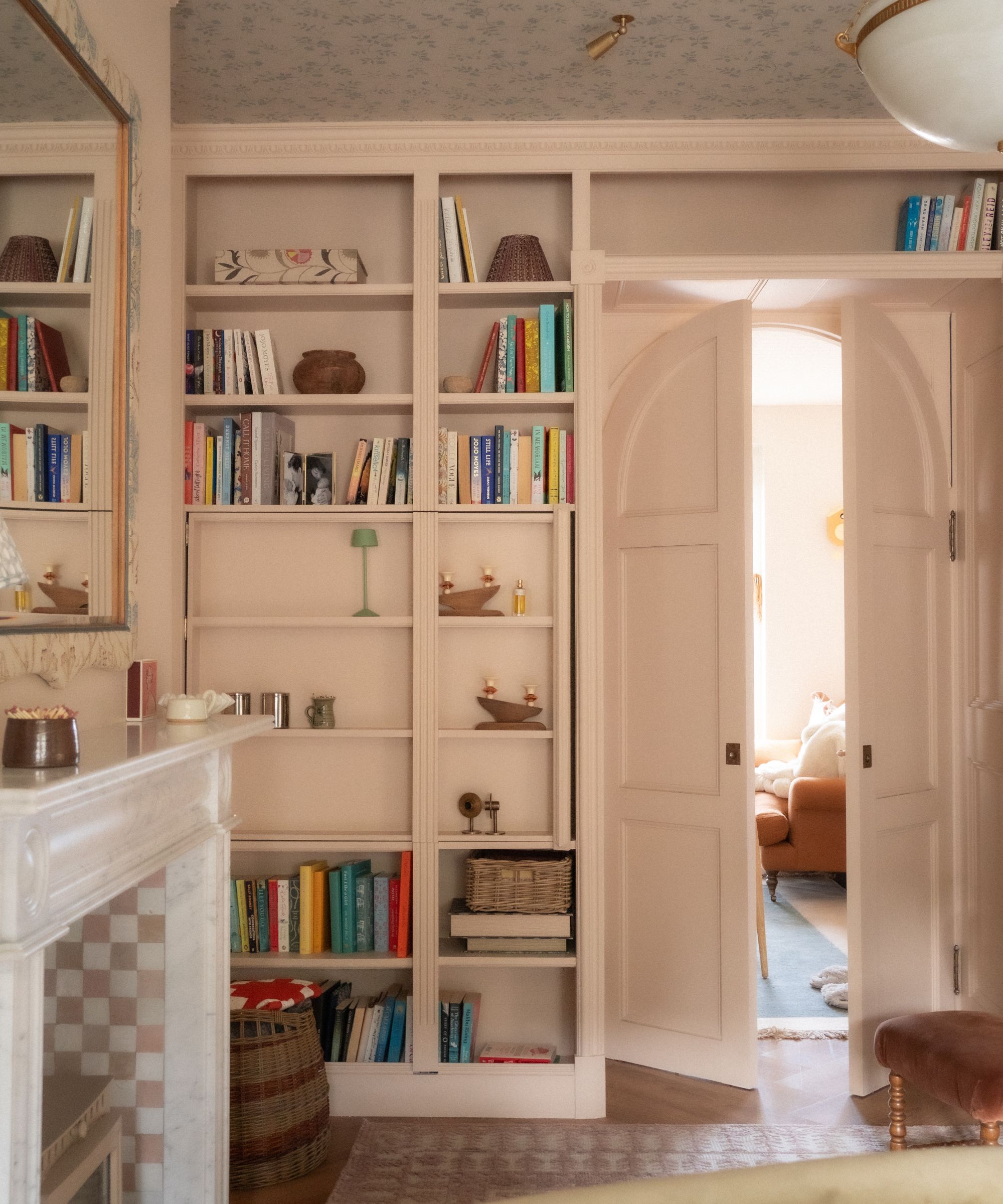 Living room with pale pink painted bookshelves and an arched doorway, shelves decorated with books and trinkets, a marble fireplace with checkerboard pink tiles inside, and a mirror above it