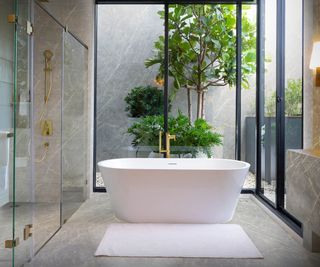 bathroom with grey marble flooring, walls, large walk in shower with glass screens, white freestanding bath and black sliding patio door leading out onto internal courtyard