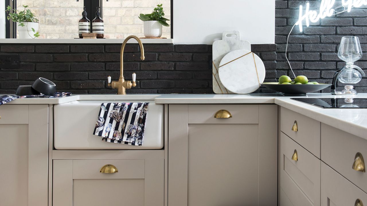 Butler sink in a beige shaker style kitchen with brass accents and a black brick wall