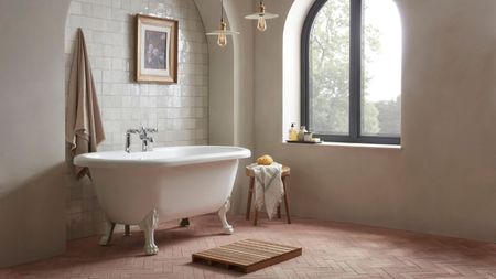 A white roll top, claw foot bathtub in a small tiled alcove in a large white painted bathroom, beside an arched window. Pink herringbone tile floor. A wooden bathmat in front of the bath. 