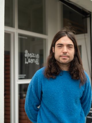 A young man wearing a blue jumper stands with his arms crossed behind his back in front of a gallery space whose sign reads "Spazio Leone" in curvilinear, whimsical white characters.