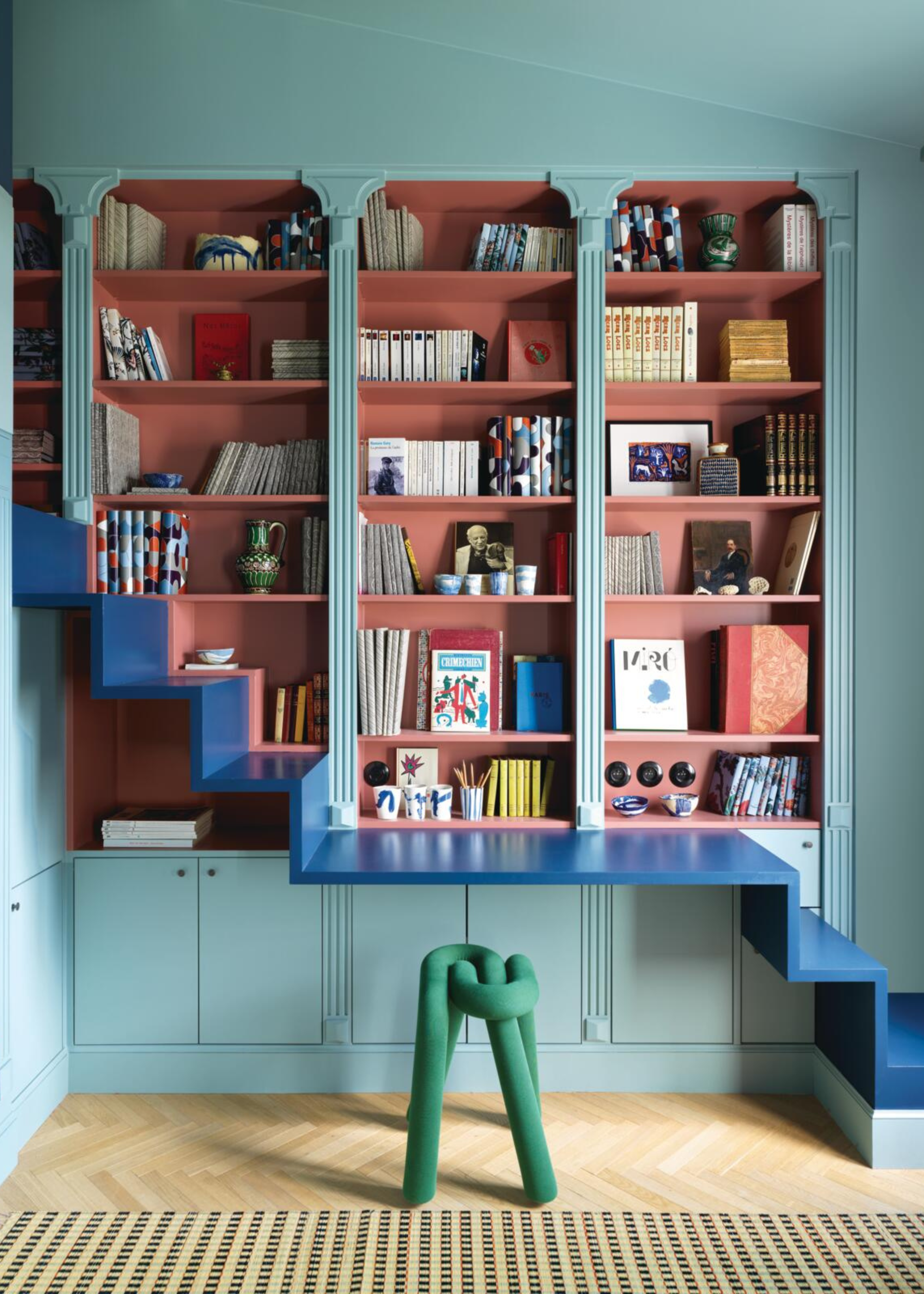 book shelving painted in a duck egg blue on the outside and terracotta on the inside with a modern blue staircase or laddering going up the side in front of it with a modern green stool in front