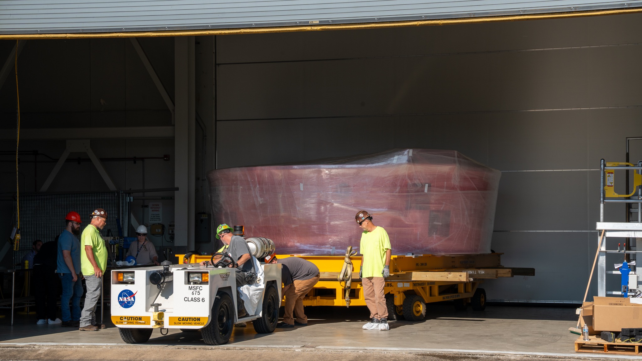 A large cylinder wrapped in plastic rests on a yellow metal frame as highlighter shirt men stand around.