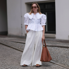 woman wearing white linen pants and a white embroidered top with a brown bag and brown shoes. 