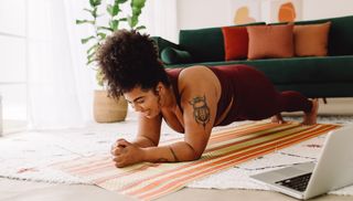 Woman holds plank position in a living room. There is a laptop to her side and a green sofa behind her.