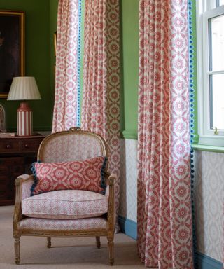 curtains with a red print and blue bobble trim against vibrant green walls with a traditional chair upholstered in a small print