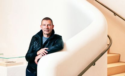 Tomas Maier, wearing black leather jacket, blue t-shirt and jeans, leaning against a curved cream coloured wall, metal hand rails and wooden steps, glass exhibition stand to the left 
