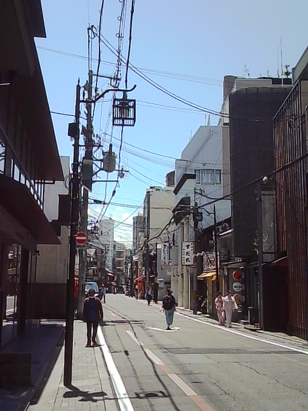 Quiet Japanese city street with pedestrians, overhead wires, and mixed modern buildings.