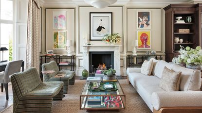 Luxe cream living room with hardwood floors and rug with paneled walls and bookshelves. There is a cream teddy four-seater sofa, and two single striped green chairs around a brass and glass coffee table. Floor-to-ceiling cabinets are built in, along with full height curtains, giving it a luxe look