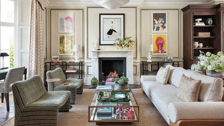 Luxe cream living room with hardwood floors and rug with paneled walls and bookshelves. There is a cream teddy four-seater sofa, and two single striped green chairs around a brass and glass coffee table. Floor-to-ceiling cabinets are built in, along with full height curtains, giving it a luxe look
