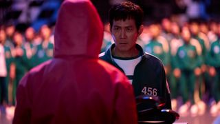 Lee Jung-jae as Seong Gi-hun, stands in front of a red-suited guard as a crowd of players in green tracksuits stand in the back, in Squid Game S2