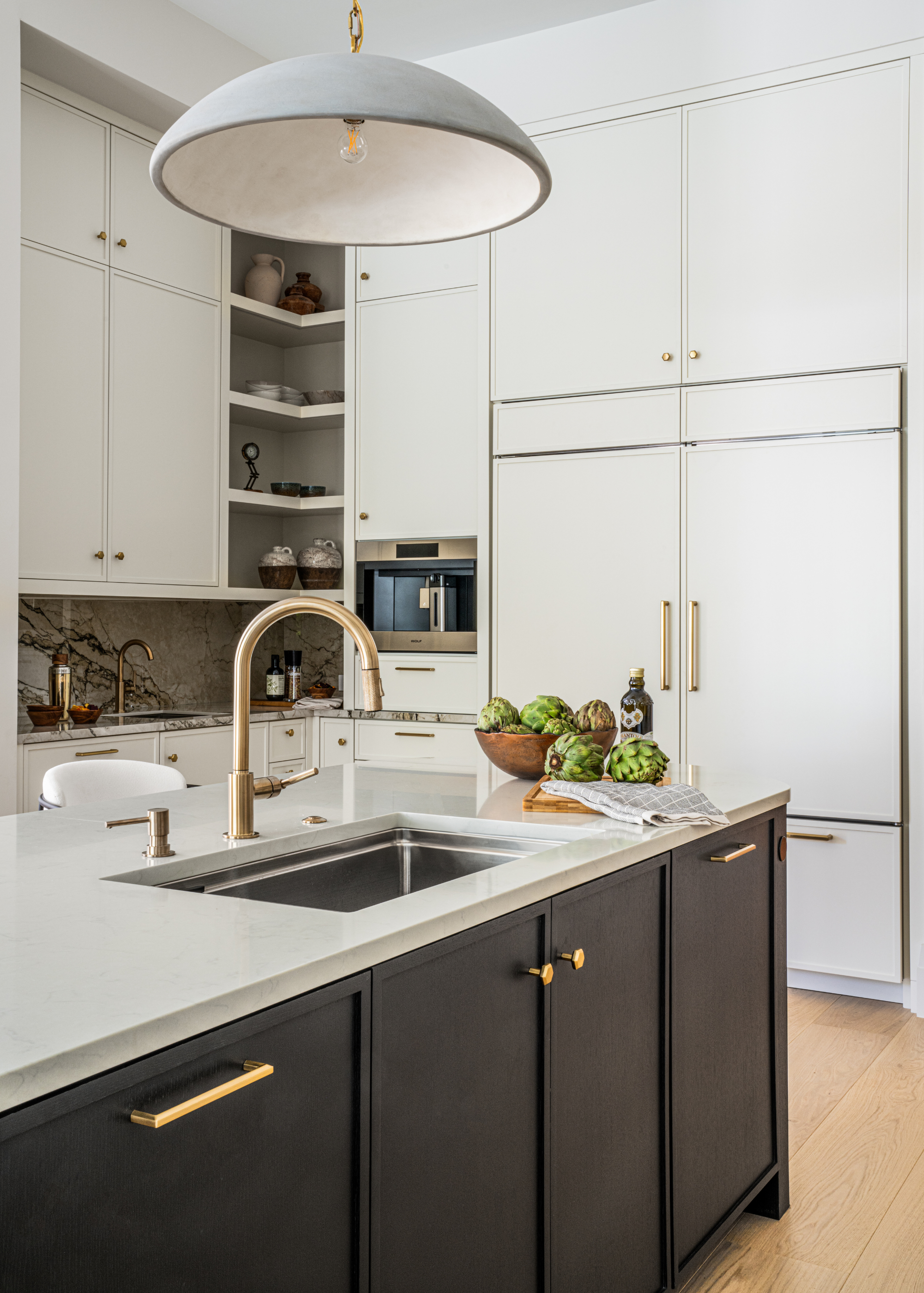 A white kitchen with gold hardware, a wood island with a stone countertop, a bowl of artichokes, a dusting cloth, and an overhead pendant lamp