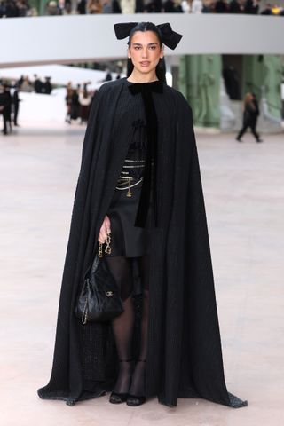 A photo of Dua Lipa wearing a cape accessorized with a black maxi hobo bag at Chanel's Haute Couture Spring/Summer 2025 show.