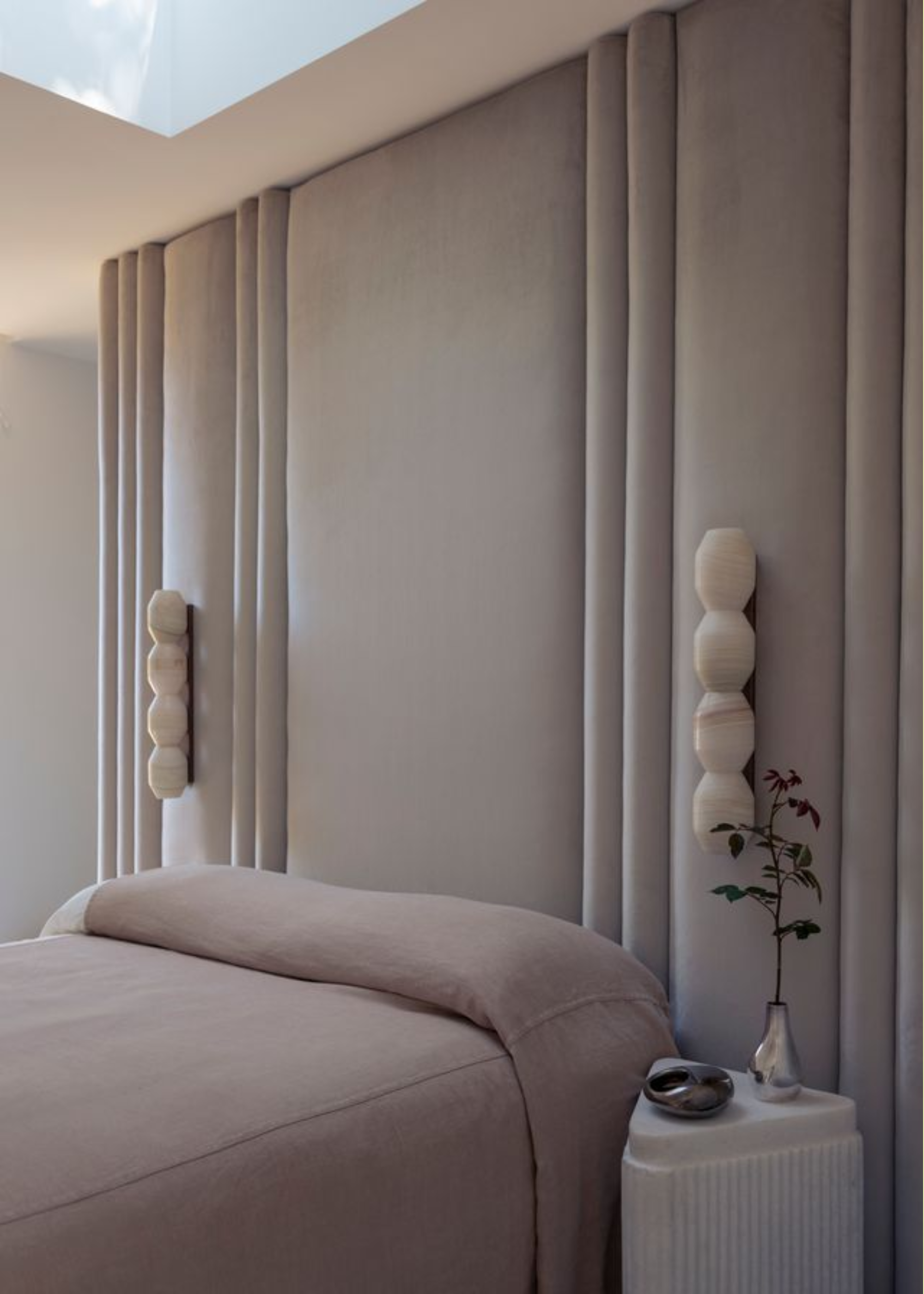 A minimalist beige bedroom with stacked wall sconces, a white stone side table with a chrome vase and a small catch-all, and a skylight