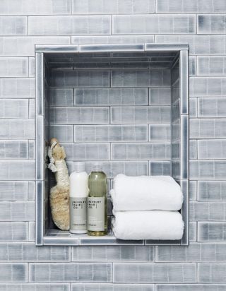 Recessed bathroom shelving with gray faded-look tiling. There are two folded fluffy white towels, shower gel and natural loofah in the recess