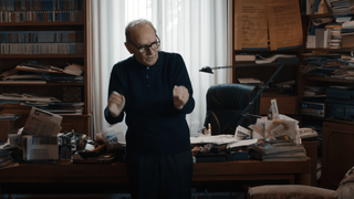 a man (ennio morricone) wearing all black clothing stands in a busy office in front of a desk, chair, and window