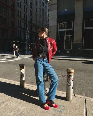 Aisha wear blue jeans and a red leather jacket as poses in front of NYC streets