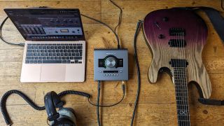A Universal Audio Apollo Twin X audio interface on a wooden floor with an electric guitar, MacBook, and a pair of studio headphones