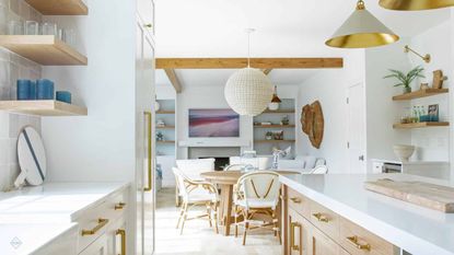 Coastal kitchen ideas are so pretty. Here is a coastal kitchen with white cabinets and wooden wall shelves to the left, a white kitchen island with wooden drawers and an oven to the right, gold and gray pendant lights