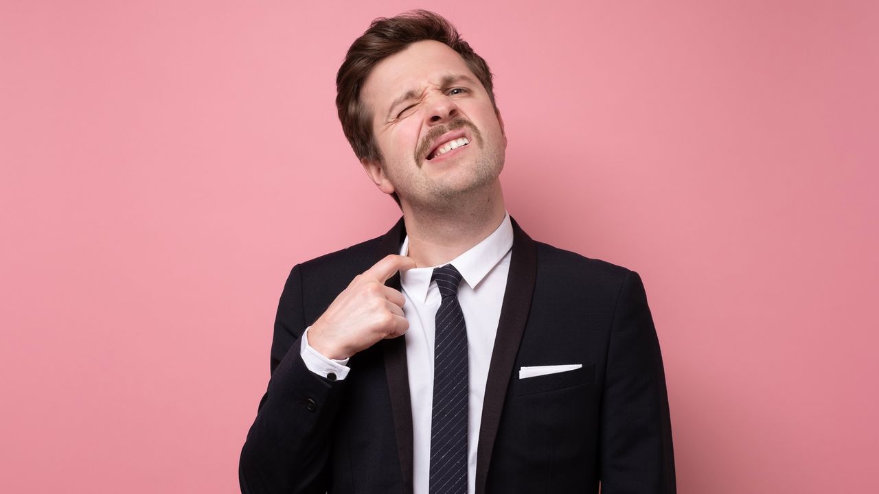 A man grimaces as he pulls a too-tight dress shirt collar away from his neck.