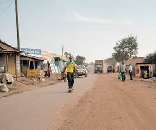 An African man riding a skateboard down a road through a village.
