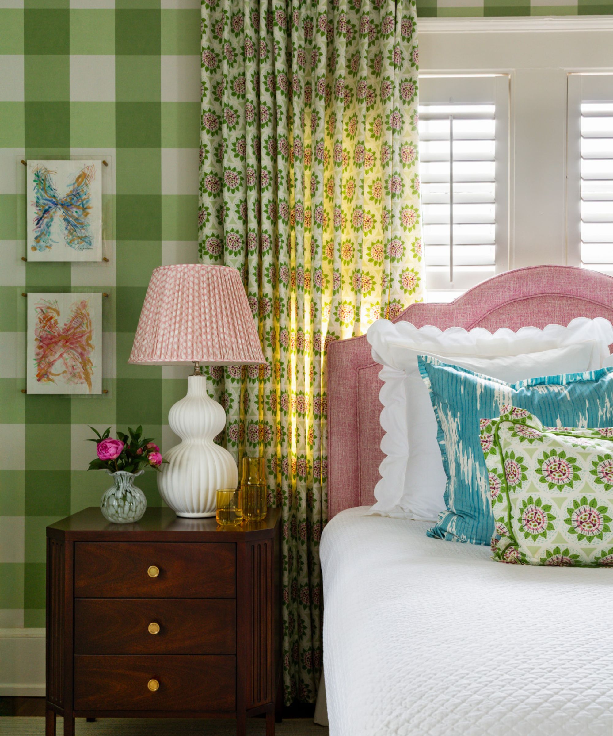 A bedroom with large-scale green gingham wallpaper, pink and green patterned curtains, white plantation shutters, a bubblegum pink headboard, white bedding, and blue and green cushions.