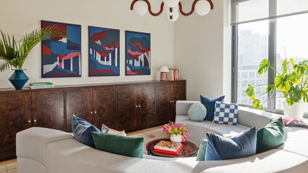 Living with dark burl wood low cabinet stretching from wall to wall, triptych of blue, red and white artworks above, pale grey curved C-shape sofa and blue and teal cushions