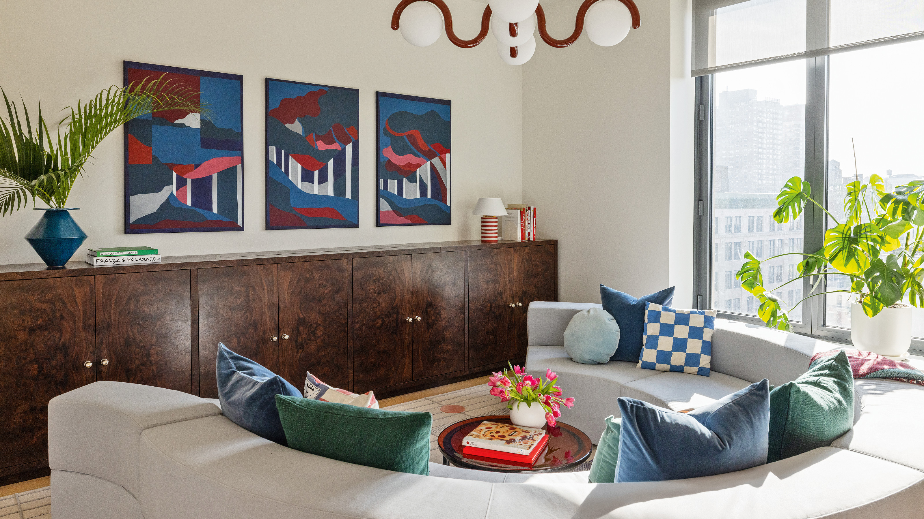 Living with dark burl wood low cabinet stretching from wall to wall, triptych of blue, red and white artworks above, pale grey curved C-shape sofa and blue and teal cushions