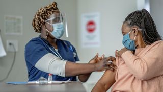 A woman wearing a surgical mask sits across from a health care provider in a mask and face shield who is preparing to give the woman a vaccine dose in her arm