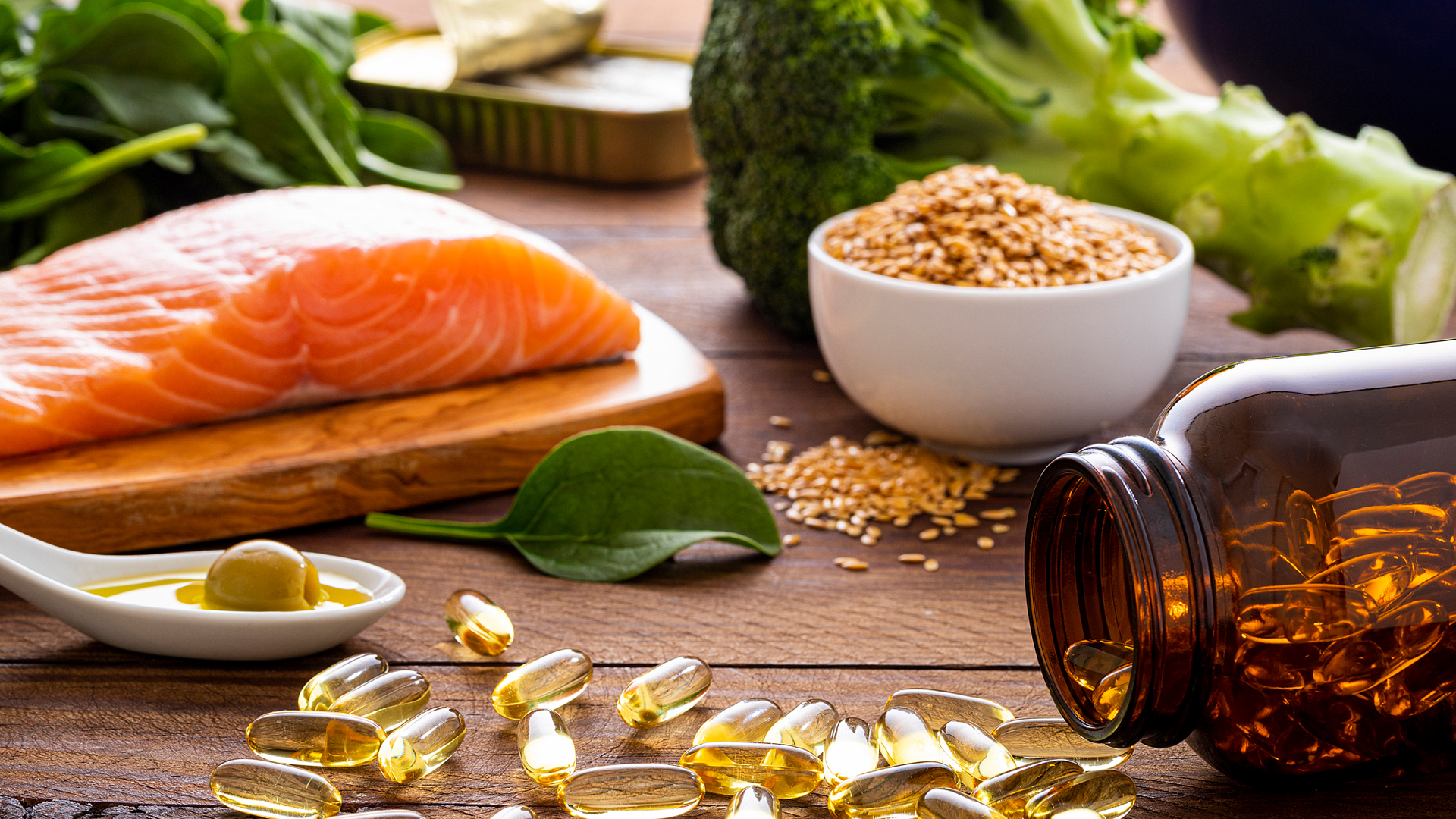 Stock image of sources of polyunsaturated fats: a filet of salmon, bowl of seeds, and spilled bottle of fish oil, all next to one another on a wood surface.