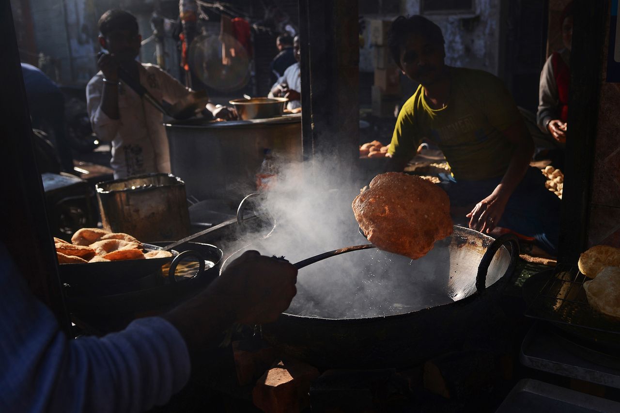 A vendor lifts a large, fried bread from a bubbling pot, surrounded by steam and other cooking elements in a bustling street food setting