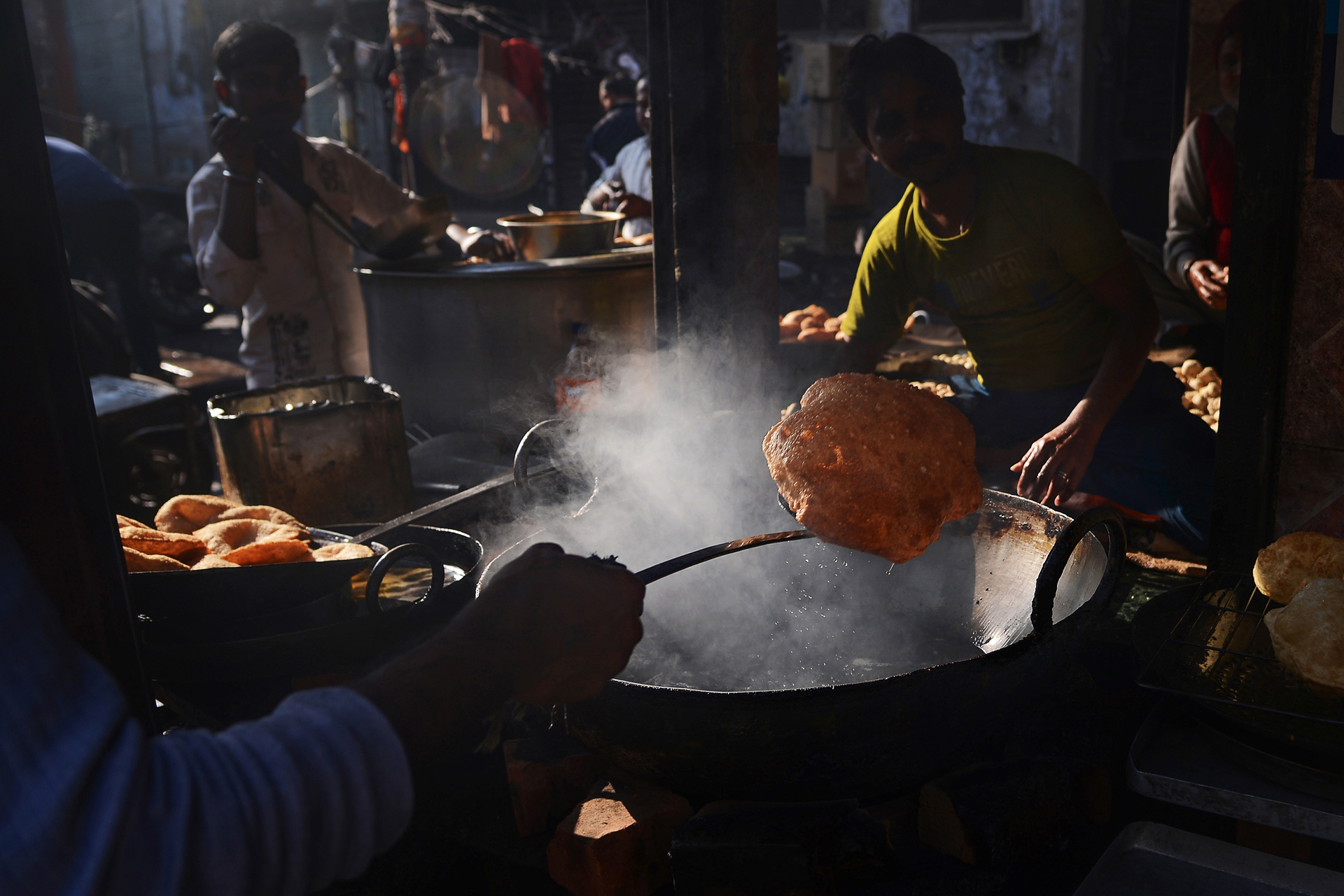 A vendor lifts a large, fried bread from a bubbling pot, surrounded by steam and other cooking elements in a bustling street food setting
