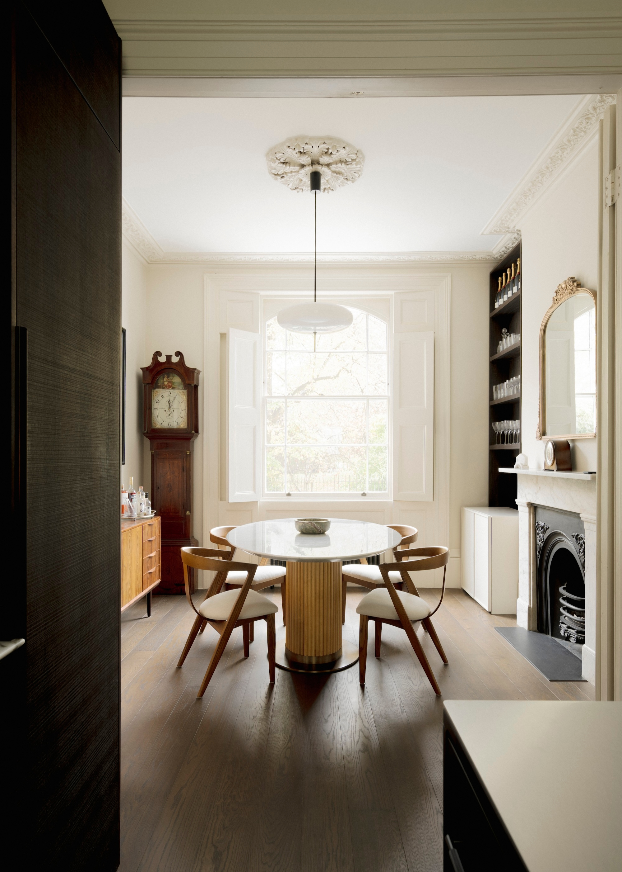 A dining room with a round glass-topped table with floor chairs around it and a pendant light hanging above in front of a fireplace with a grandfather floor clock in the corner and dark wood flooring
