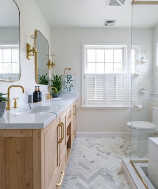 A white bathroom with two gold curved mirrors and wall sconces, a light brown sink unit with gold finishes and a marble surface, a white toilet, and a clear glass shower