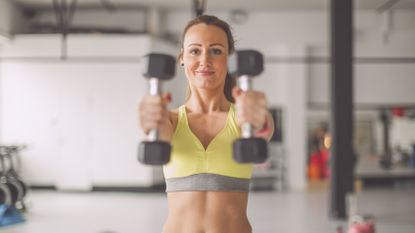 Woman smiling and holding light weight dumbbells out in front of her in a gym