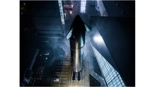 a large silver rocket rolls down a road at night, passing close to a lit-up building