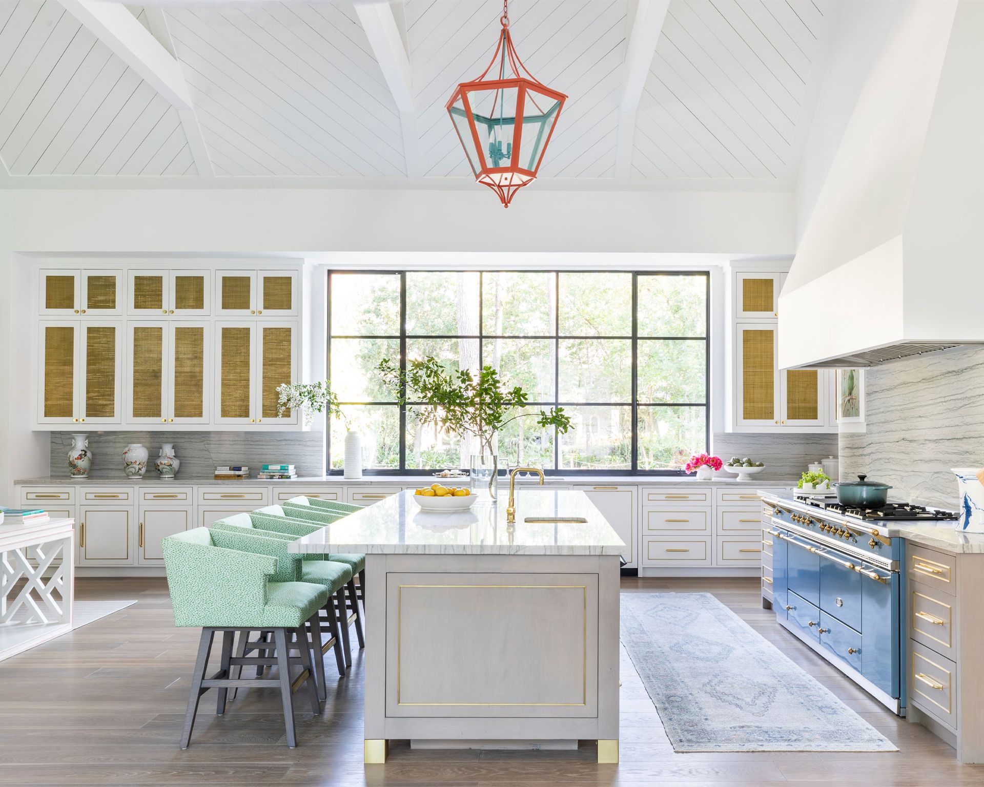 Large kitchen with white wooden paneled ceiling, orange metal and glass pendant, large kitchen island with seating, rug on floor between island and stove, white painted walls, gray cabinetry