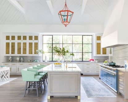 Large kitchen with white wooden paneled ceiling, orange metal and glass pendant, large kitchen island with seating, rug on floor between island and stove, white painted walls, gray cabinetry