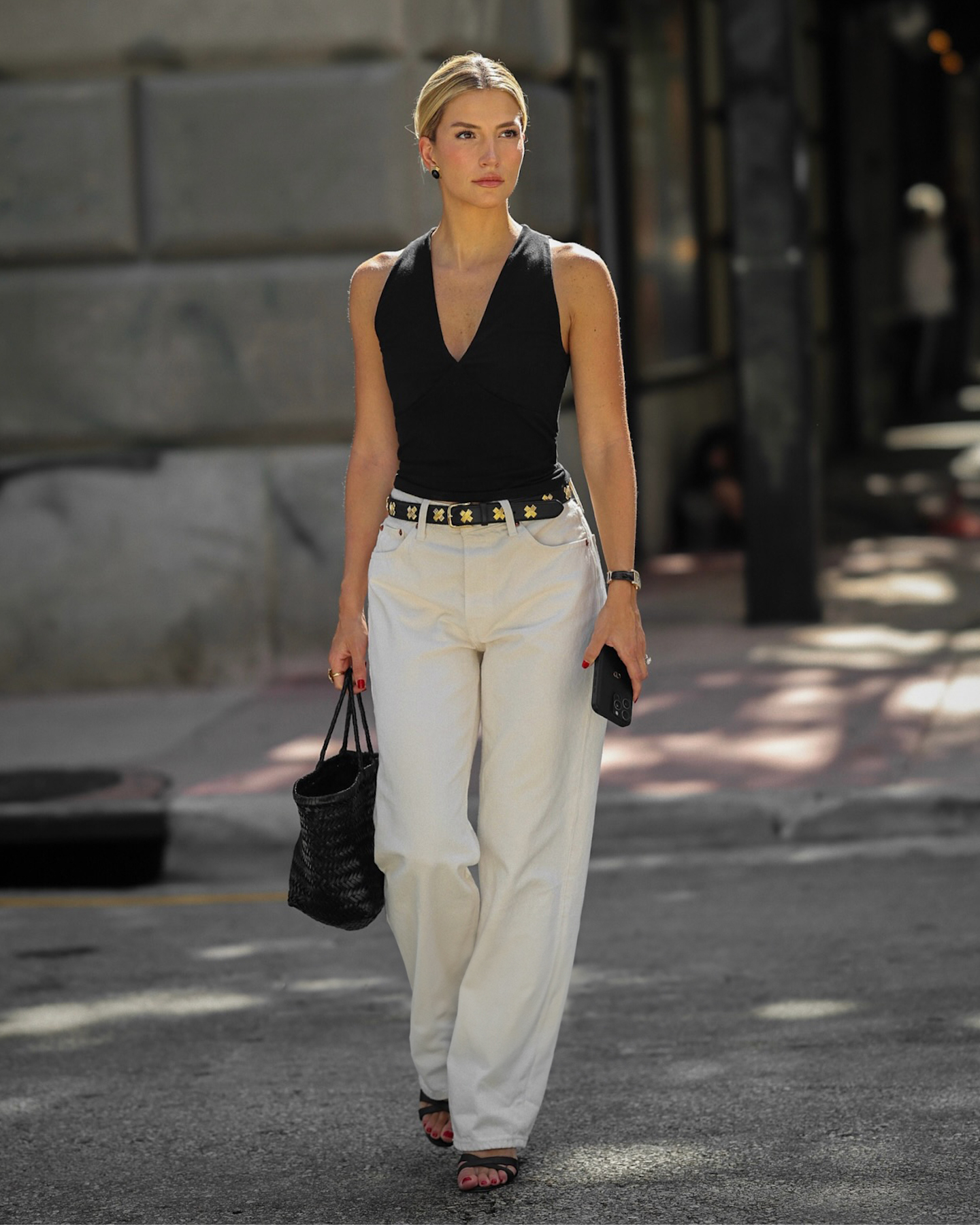 style influencer Cass Dimicco crossing a street wearing a black w-neck top, studded Aureum Collective belt, ivory jeans, a black woven tote bag, and black strappy sandals.