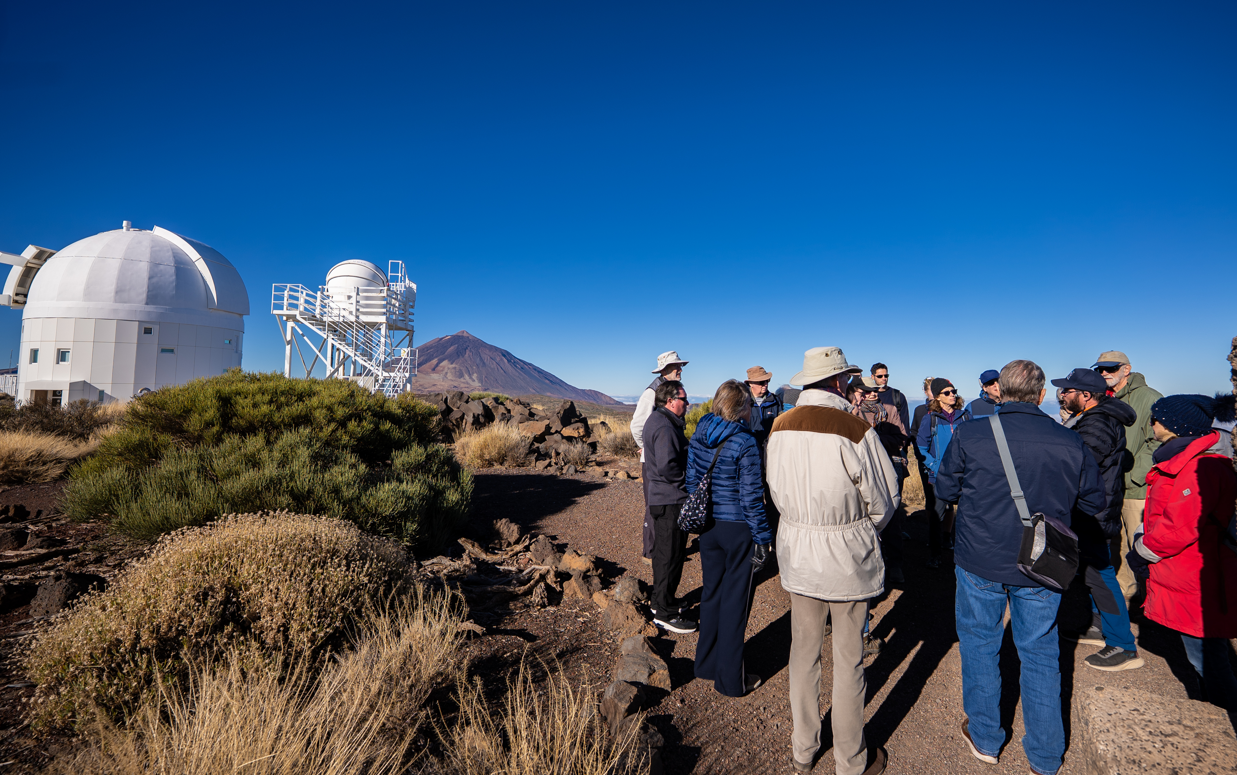 a group of people stand listening to a guide, there is a telescope dome on the left and a volcano in the distance.